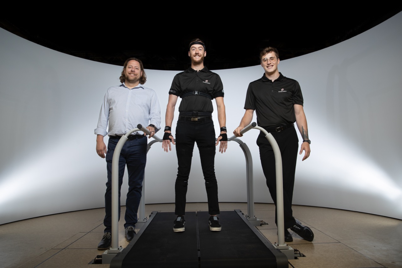 Three people, one on large treadmill with sensors placed on hands and head.