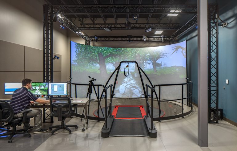 One person sits at desk with large treadmill and large curved screen displaying trees and a stone path