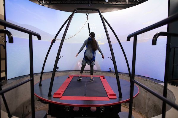 Person in harness on large treadmill in front of very large curved screen displaying coastline