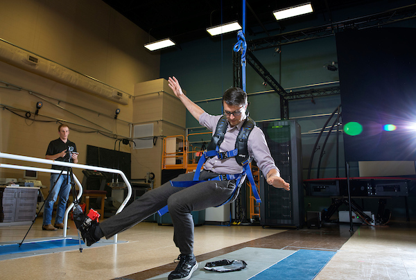 Person strapped into harness, demonstrating bending of left knee while second person stands by to witness and record