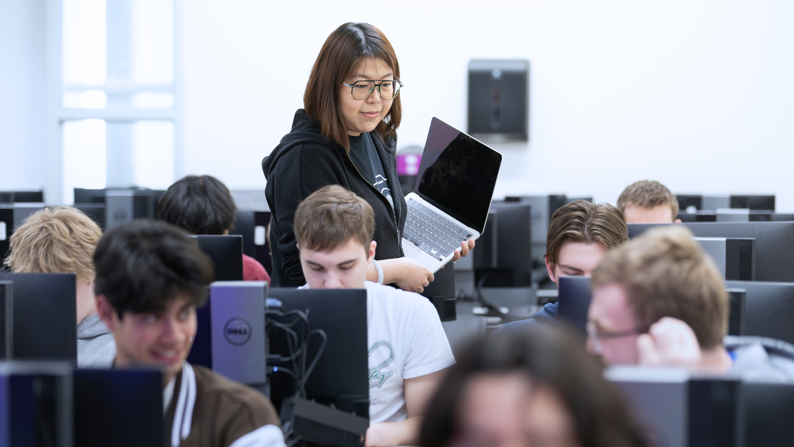 A woman with shoulder-length brown hair and glasses, wearing a black hoodie, stands in a computer lab holding a laptop. As the instructor of the computer science course, she is moving between rows of computer workstations where students are seated. The students, visible from behind or in profile, are focused on their computer screens in the university computer lab with multiple desktop monitors visible throughout the space. Large windows with natural light can be seen in the background.
