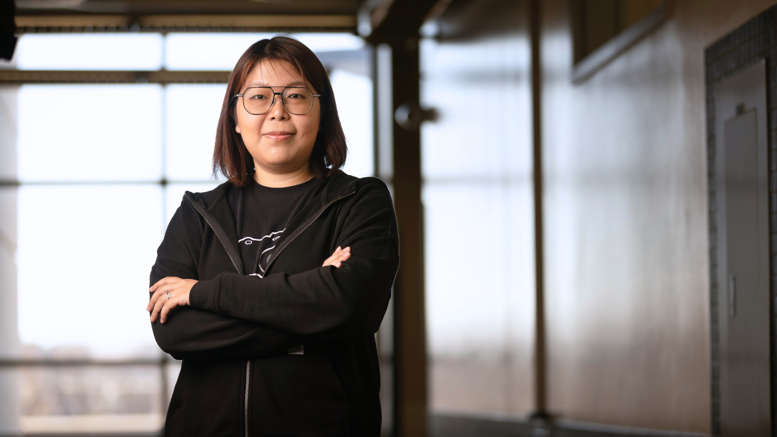 A woman with shoulder-length brown hair and round glasses stands with her arms crossed, looking confidently at the camera. She is wearing a black hoodie over a black t-shirt with a simple white design on it. The background shows a modern interior space with large windows allowing natural light to filter in. The setting is an academic building with minimal architectural elements visible.