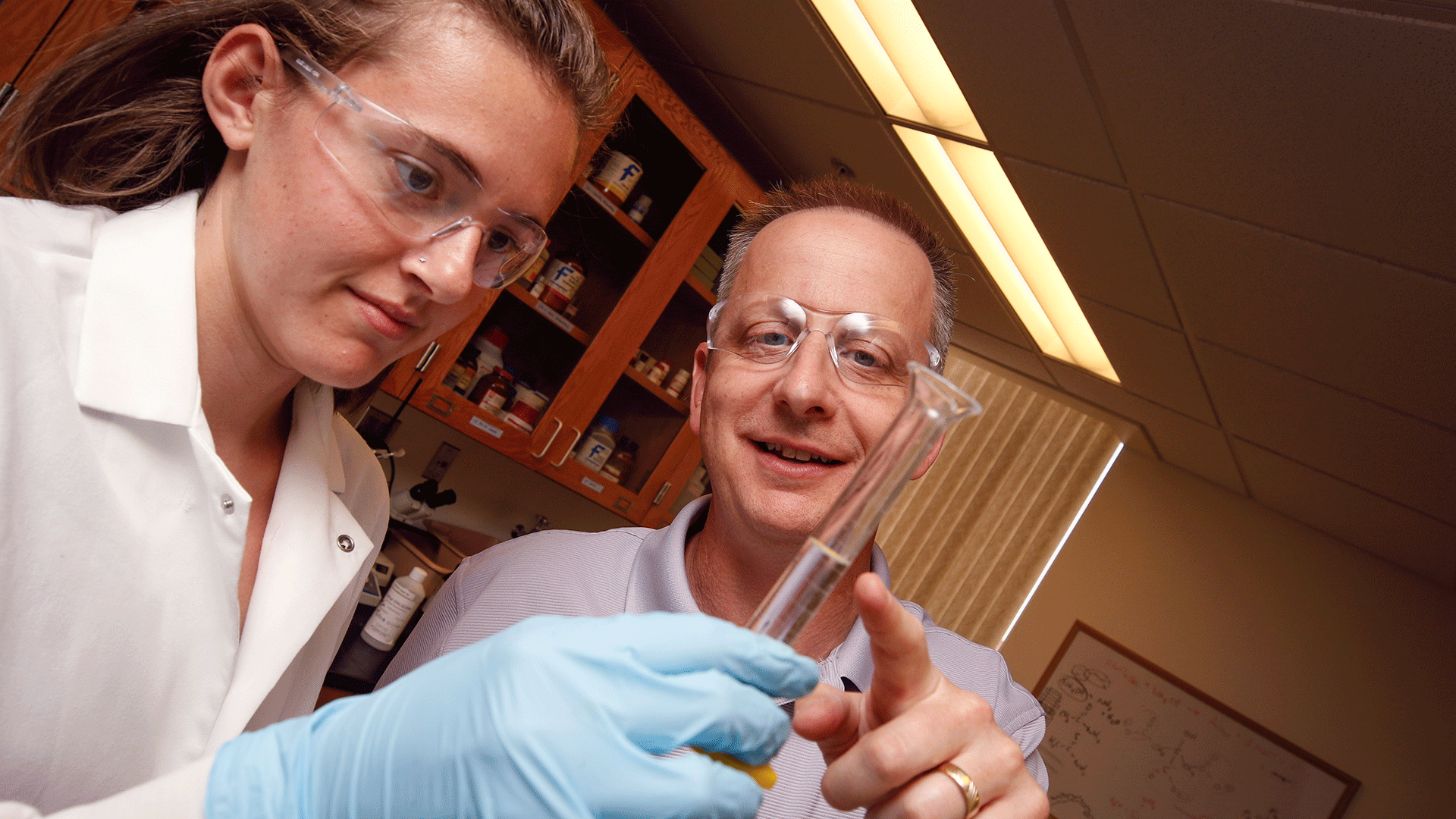 female in white lab coat holding beaker while man points to contents