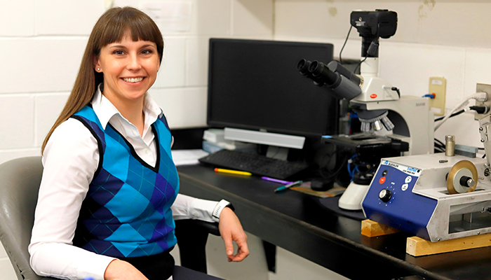 female scientist seated at counter in front of microscope and monitor, smiling at the camera