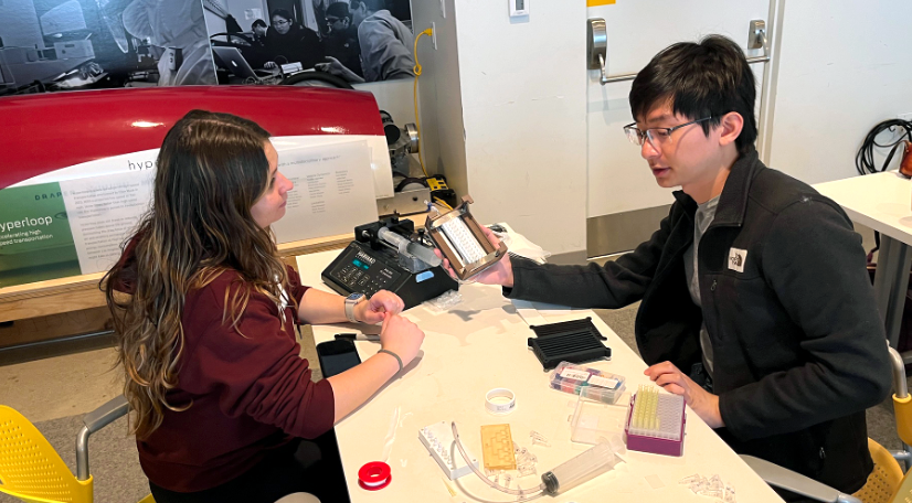 Two individuals sit at a table in a lab, examining scientific equipment. The table displays various lab items, including a pipette and test tubes. In the background, there is a large poster and lab equipment.