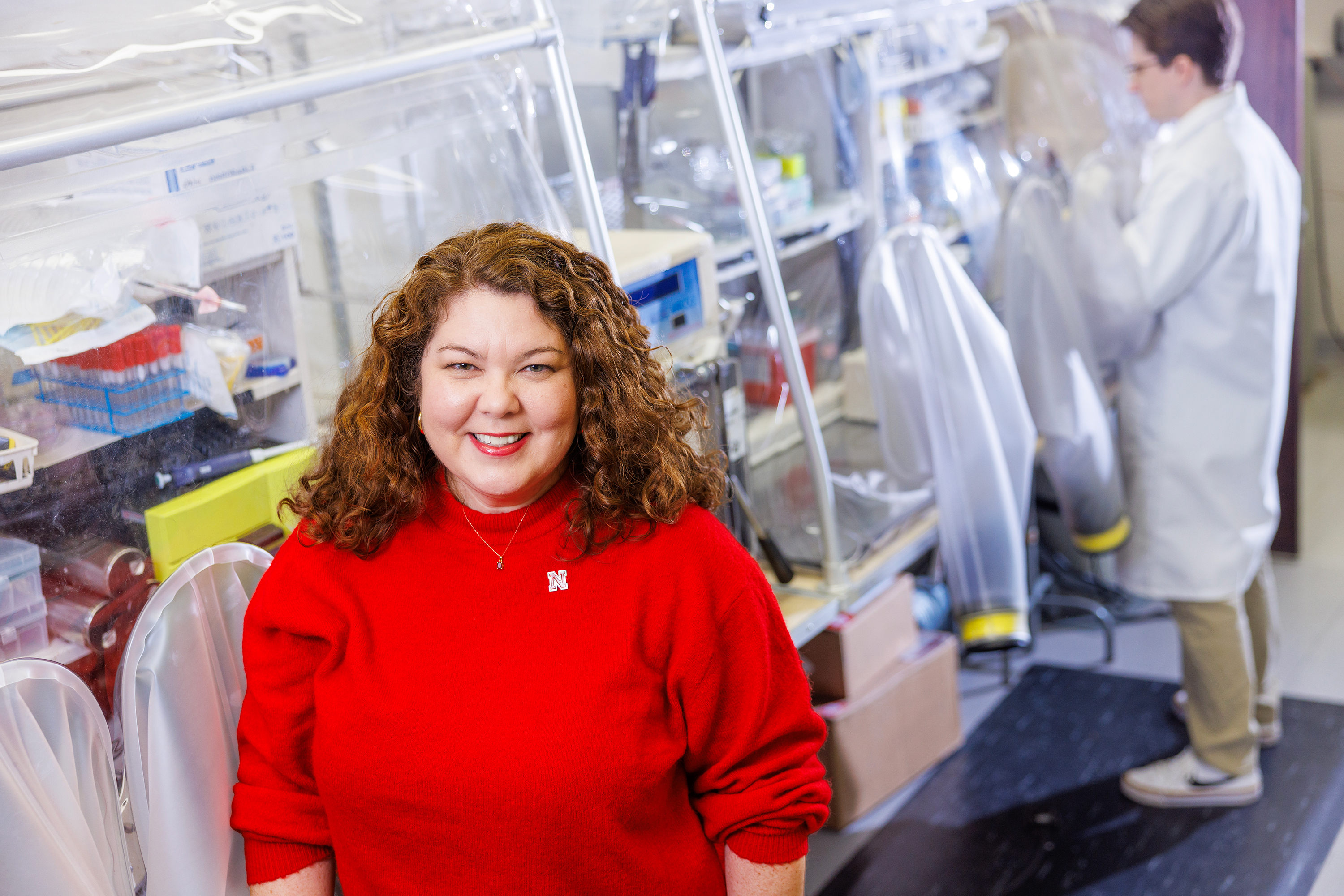 Female researchers in bright red sweater with UNL pin smiles at camera. In the background scientist in a lab coat at the bench.