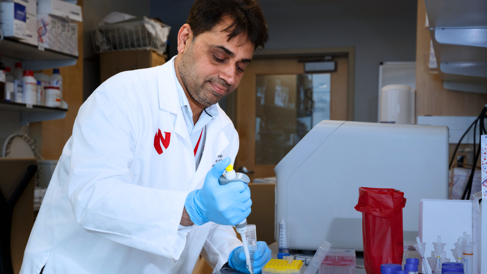 A laboratory scientist wearing a white lab coat with a red logo and blue nitrile gloves is focused on pipetting liquid into a small tube. The researcher is working at a lab bench surrounded by scientific equipment, red biohazard waste bag, and various laboratory consumables like test tubes and pipette tips. The laboratory setting includes shelves with reagent bottles and supplies visible in the background.