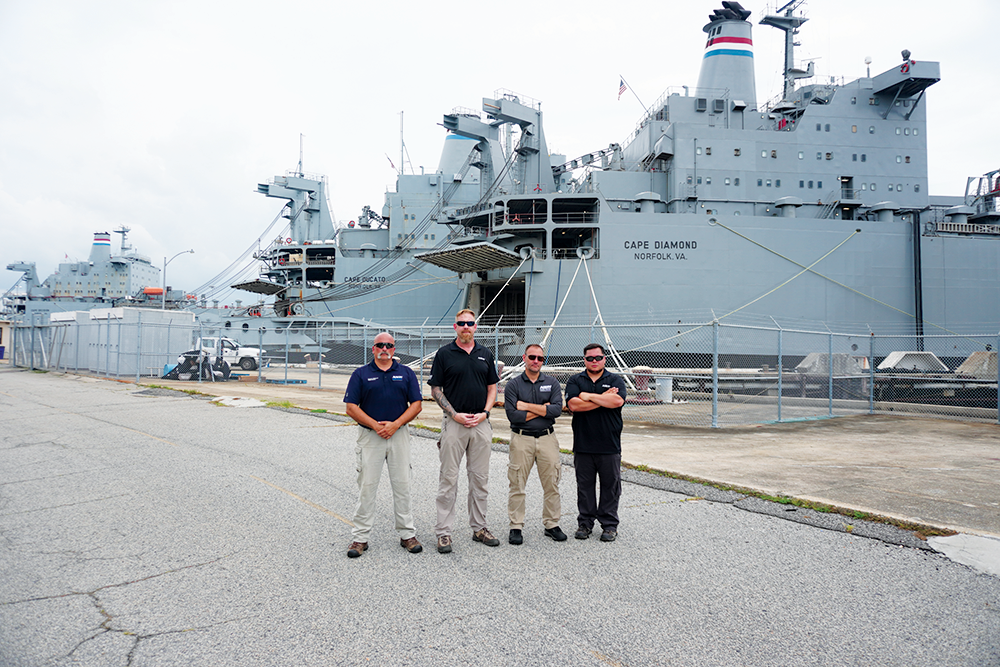 Four men standing with arms crossed in front of huge naval ship