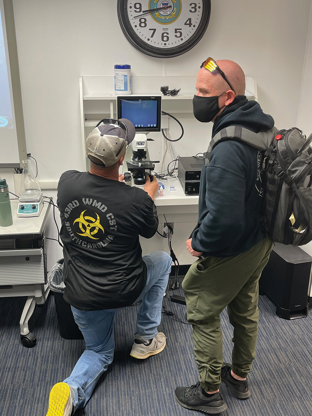 Man kneeling in front of microscope looking at screen, another man standing ot the right viewing