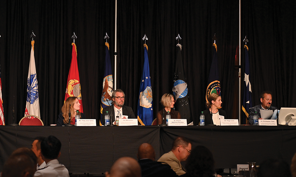 panel of five speakers seated behind table with military flags in background