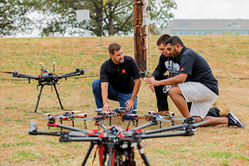 Two individuals kneeling on a grassy field inspecting a large multi-rotor drone, with another drone and outdoor equipment visible in the background.