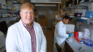 A laboratory workspace where a person in a white lab coat with a purple/pink button-up shirt is smiling at the camera in the foreground. Another person in a white lab coat works at a lab bench in the background. The lab contains various scientific equipment, shelves stocked with boxse and laboratory supplies. The setting is a scientific research and medical laboratory with typical equipment like test tubes, racks, and packaging materials visible on the counters and shelves.