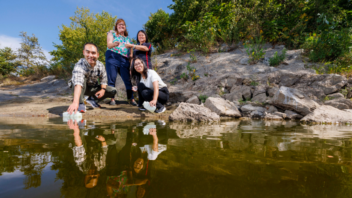 Four University of Nebraska researchers collect water samples at the edge of a shallow outdoor waterway. Two team members crouch near the water's edge holding collection bottles while two colleagues stand behind them. The group's reflections are visible on the calm water surface. Rocky outcroppings and dense green vegetation line the bank behind them under a clear blue sky.