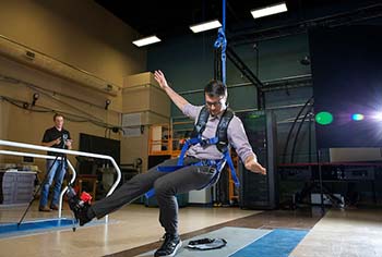 Person strapped into harness, demonstrating bending of left knee while second person stands by to witness and record