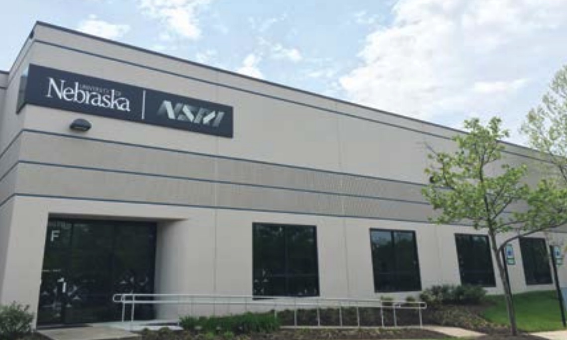 “Exterior view of the NSRI facility in a light‑colored commercial building, featuring a sign with the University of Nebraska and NSRI logos above the entrance. The building has large windows, a ramp leading to the door, and landscaping with small trees and shrubs.