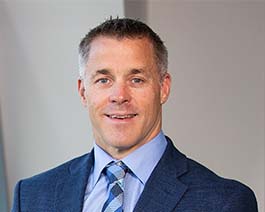 Professional headshot of Carlstrom in a blue suit and patterned tie, smiling against a neutral office background