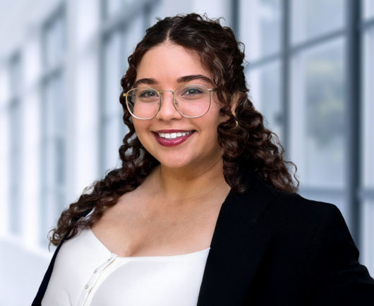 Professional headshot of Cruz-Rodriguez with curly hair and glasses, wearing a black blazer and light blouse in a bright modern office setting