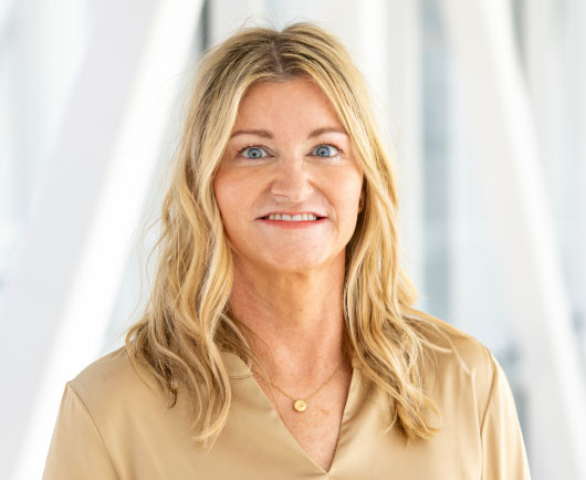 Professional headshot of a smiling Ravnholdt with blonde hair, wearing a beige blouse in a bright, modern office environment