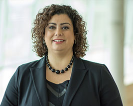 Professional headshot of April Shea with curly hair, wearing a black blazer and statement necklace in a modern office setting