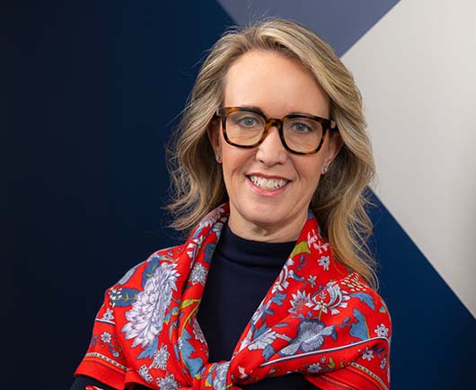Professional headshot of Anne Barnes wearing glasses and a red patterned scarf, posed against a modern geometric background