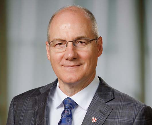 Professional headshot of Bayles with glasses, wearing a gray plaid suit and blue tie in a softly lit office setting