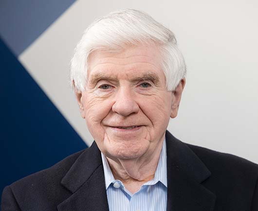 Professional headshot of Ben Nelson, wearing a gray plaid suit and blue tie in a softly lit office setting