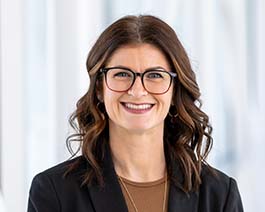 Professional headshot of Bensman with wavy brown hair and glasses, wearing a black blazer and brown top in a bright office setting