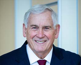 Professional headshot of Bob Hinson with gray hair, wearing a dark suit and red tie in a bright office environment
