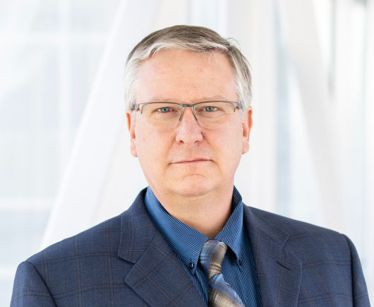 Professional headshot of a serious-looking man with glasses, wearing a blue suit and patterned tie in a bright, modern office setting