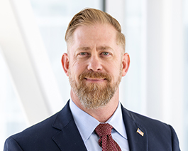 Professional headshot Dan Polanski, wearing a navy suit and red tie with an American flag pin in a bright office setting