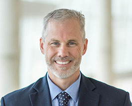 Professional headshot of Dave Roberts with short gray hair and a trimmed beard, wearing a navy suit and polka dot tie in a bright office setting
