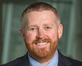 Professional headshot of Dillan Cunningham with a red beard, wearing a suit and tie in a modern office setting