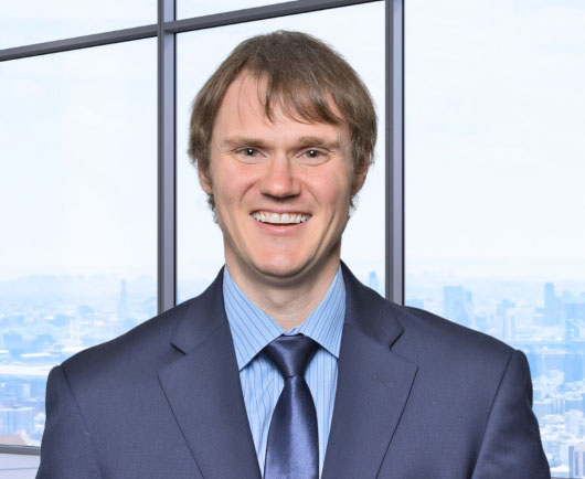 Professional headshot of Distelhorst in a blue suit and tie, standing in front of large office windows with a city skyline background