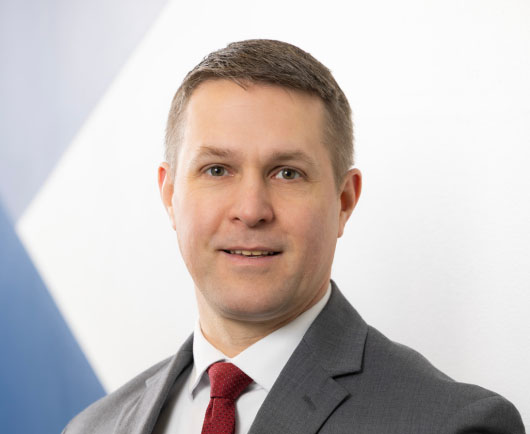 Professional headshot of Erik Johnson in a gray suit and red tie, looking at the camera against a clean, modern background