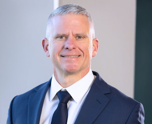 Professional headshot ofGeist with short gray hair, wearing a navy suit and tie in a bright office environment