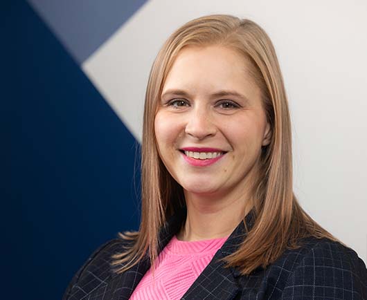 Professional headshot of Gonnerman with straight blonde hair, wearing a dark blazer and pink top against a modern geometric background