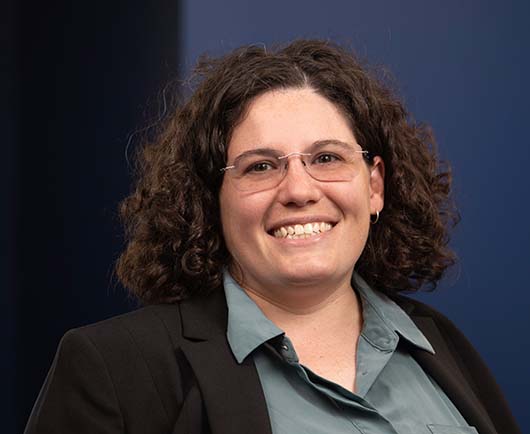 Professional headshot of Meghan Jackson with curly hair and glasses, wearing a dark blazer and green blouse against a blue studio background