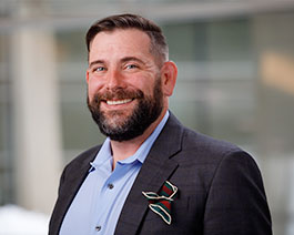 Professional headshot of John Santarpia with a beard, wearing a blazer and light blue shirt with a colorful pocket square in an outdoor office setting