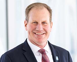 Professional headshot of Chris in a dark suit and red patterned tie, posed in a bright office setting