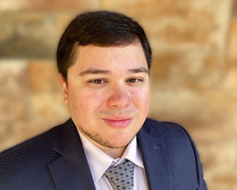 Professional headshot of Marty Sikes III with short dark hair, wearing a suit and patterned tie, posed against a warm-toned textured background