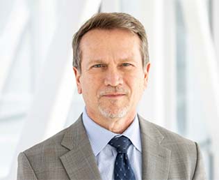 Professional headshot of Marty Sikes with short hair and a trimmed goatee, wearing a gray suit and blue tie in a bright office setting