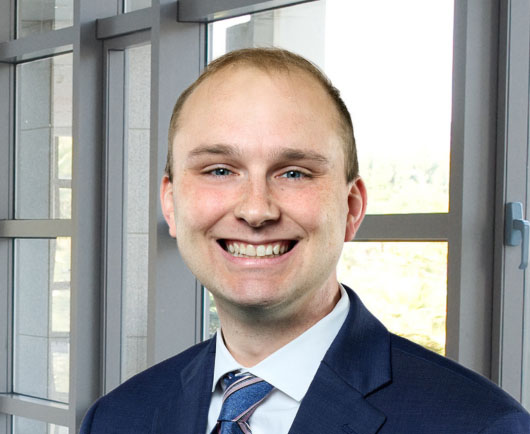 Professional headshot of Matt Sweeney in a blue suit and striped tie, standing in front of large office windows