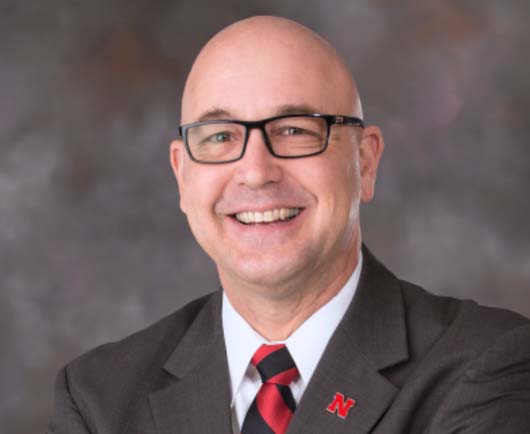 Professional headshot of Mike Boehm wearing glasses, a dark suit, and a red striped tie with a lapel pin against a neutral studio background