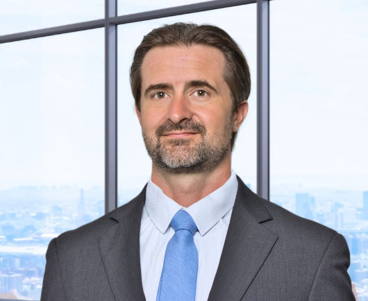 Professional headshot of Minter with dark hair and light stubble, wearing a gray suit and blue tie in front of large office windows with a city skyline background