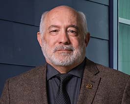 Professional headshot of Neal Woollen with a gray beard, wearing a brown blazer and black shirt and tie against a modern exterior wall background