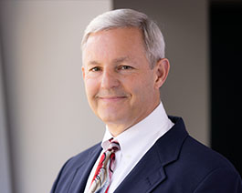 Professional headshot of Patrick Rhoads with gray hair, wearing a navy suit and patterned tie against a clean, softly lit background