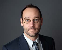 Professional headshot of Paul Brantmier with glasses and light stubble, wearing a suit and tie against a dark studio background