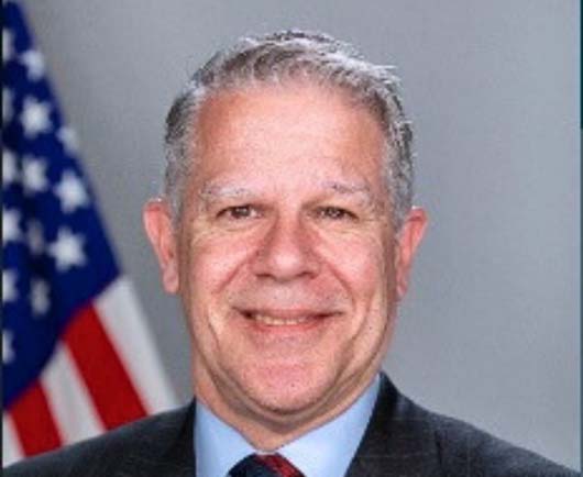Professional headshot of Paul Friedrichs with gray hair, wearing a suit and tie with an American flag in the background