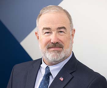 Professional headshot of Richard Evans with gray hair and beard, wearing a navy suit and tie with an American flag pin against a modern geometric background