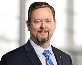 Professional headshot of Rick Arestad with short hair and a goatee, wearing a navy suit and blue striped tie in a bright office setting